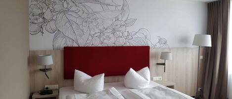 Habitación de hotel con un gran mural floral en la pared, un cabecero rojo, dos lámparas de noche y una cama bien hecha con sábanas blancas.