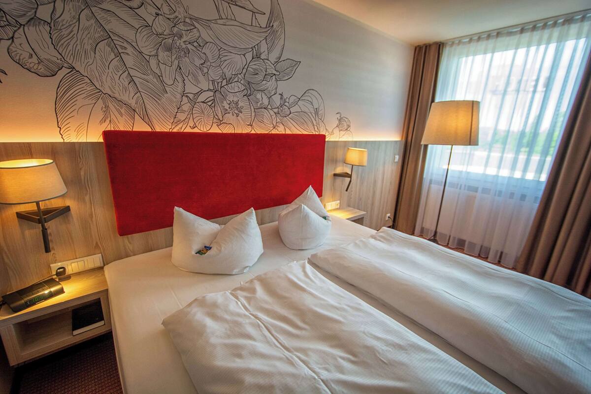 A hotel room with a large bed, two white pillows, a red headboard, a wall mural, a nightstand with a lamp, and a window with curtains.
