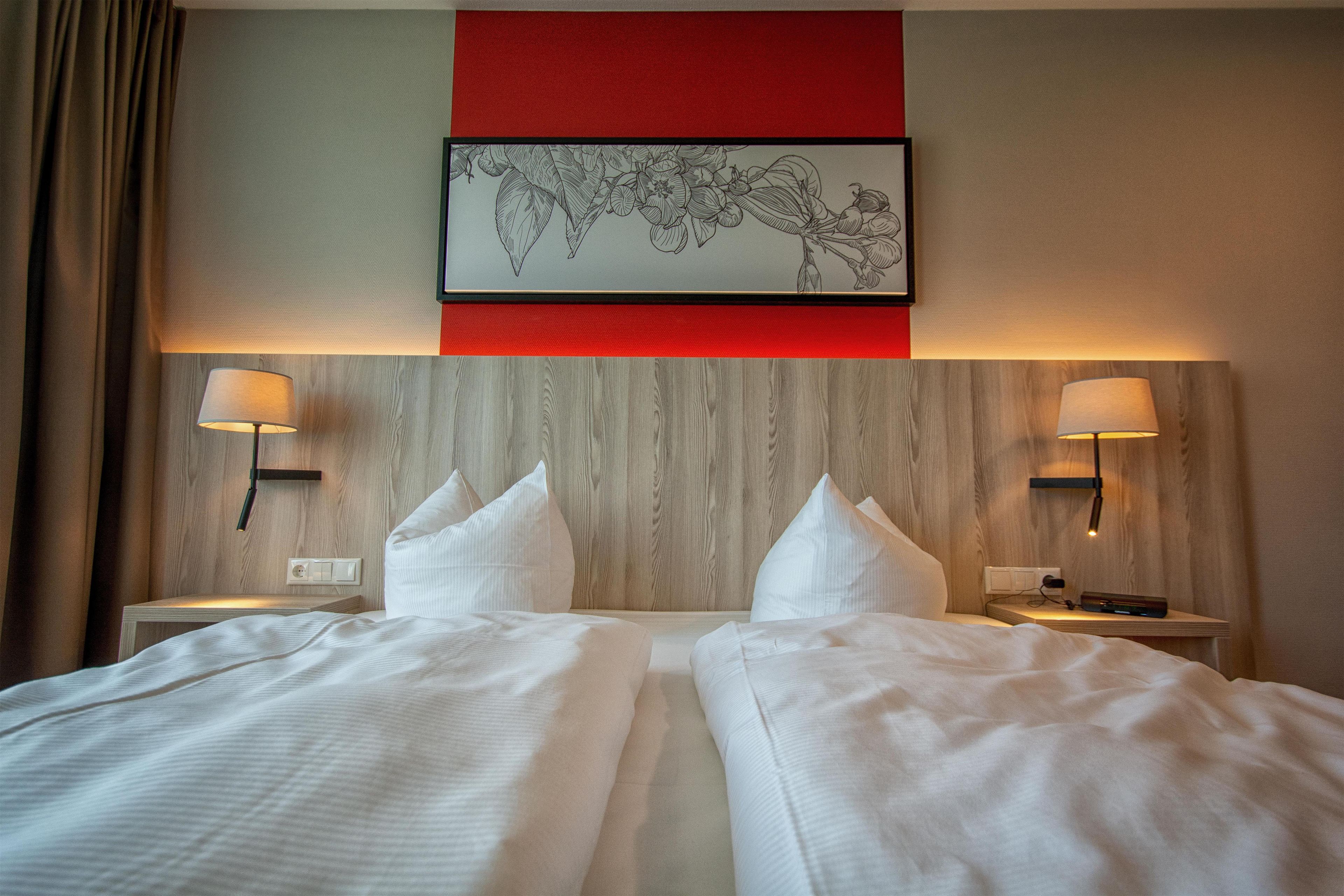 A hotel room with two beds, a red accent wall, and a framed botanical illustration.