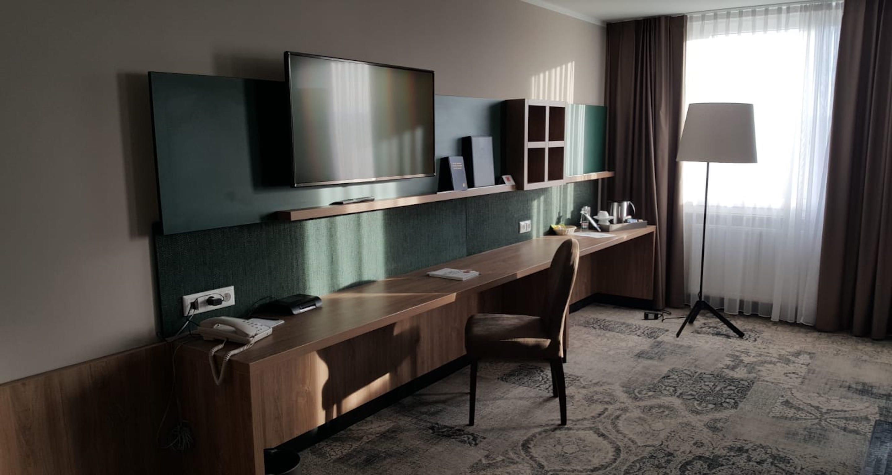 Ein Hotelzimmer mit einem langen Holztisch, einem an der Wand montierten Fernseher, einem Sessel, einer Lampe und einem gemusterten Teppich.