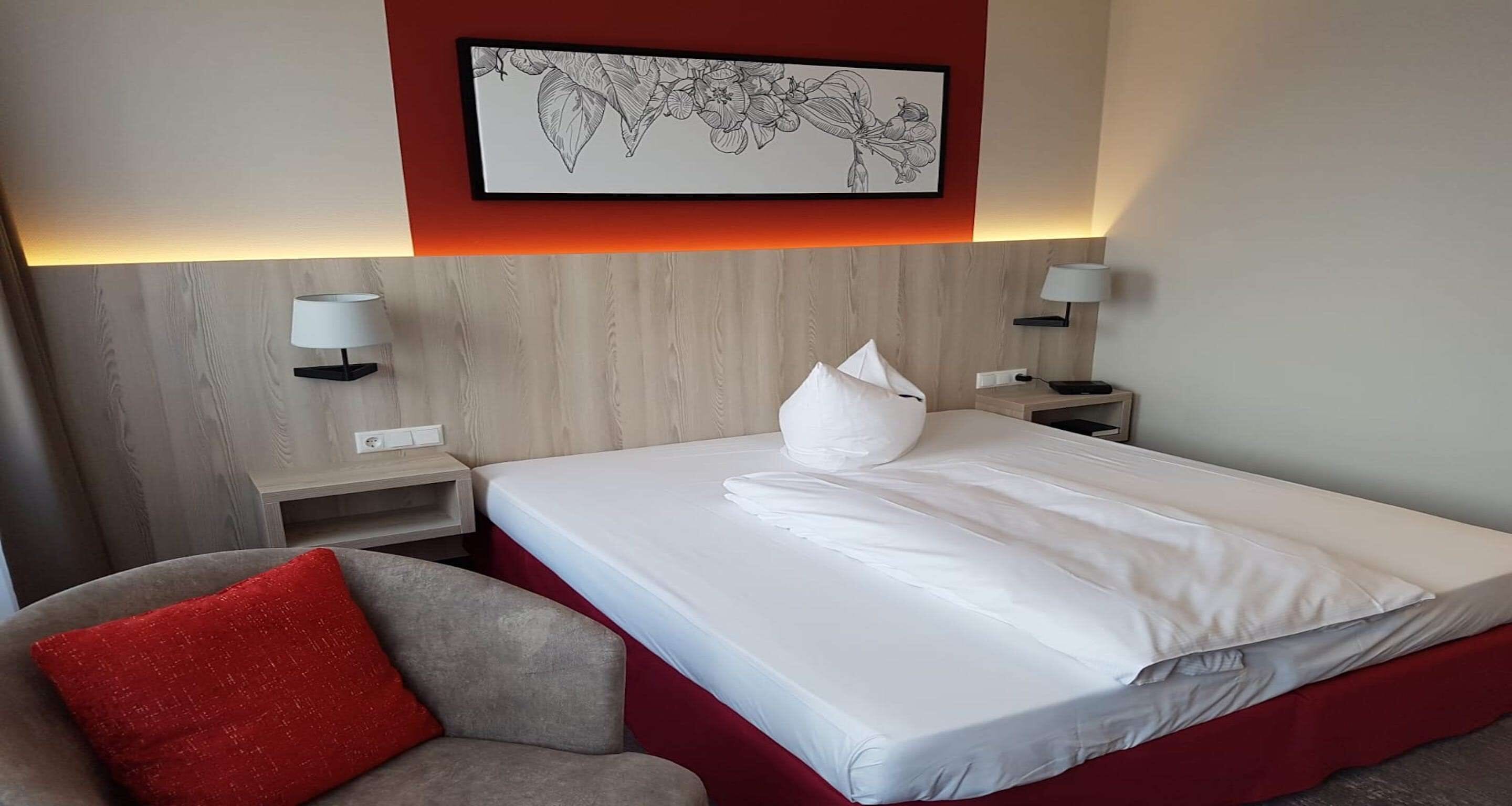 Ein Hotelzimmer mit einem Bett, einer roten Akzentwand, einer gerahmten Blumenblume, zwei Nachttischlampen und einem roten Kissen auf einem grauen Sessel.