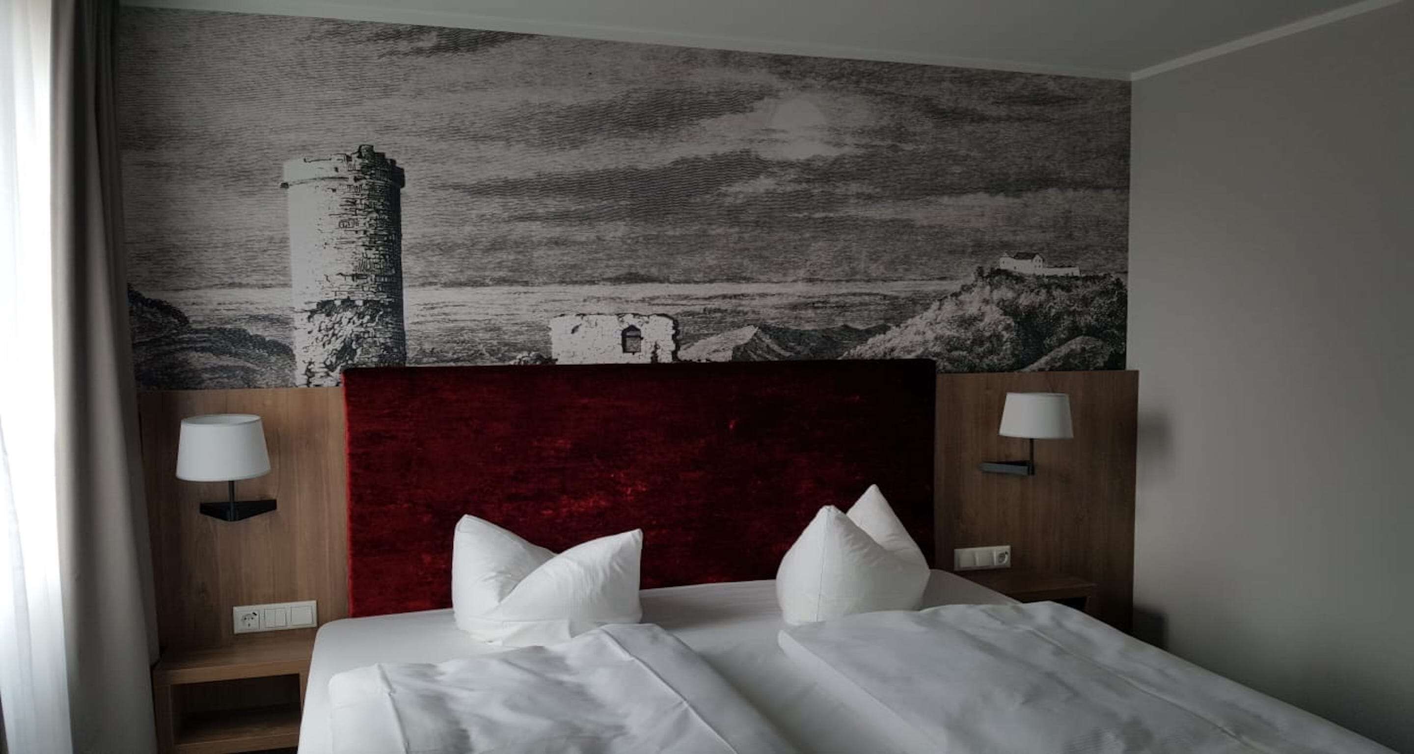 Ein Hotelzimmer mit einem Bett, zwei Nachttischlampen, einem roten Kopfteil und einer großen Wandmalerei mit einer Küstenlandschaft.