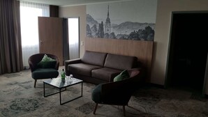 Habitación de hotel con sofá marrón, dos sillones, una mesa de centro y un cuadro de una iglesia en la pared.