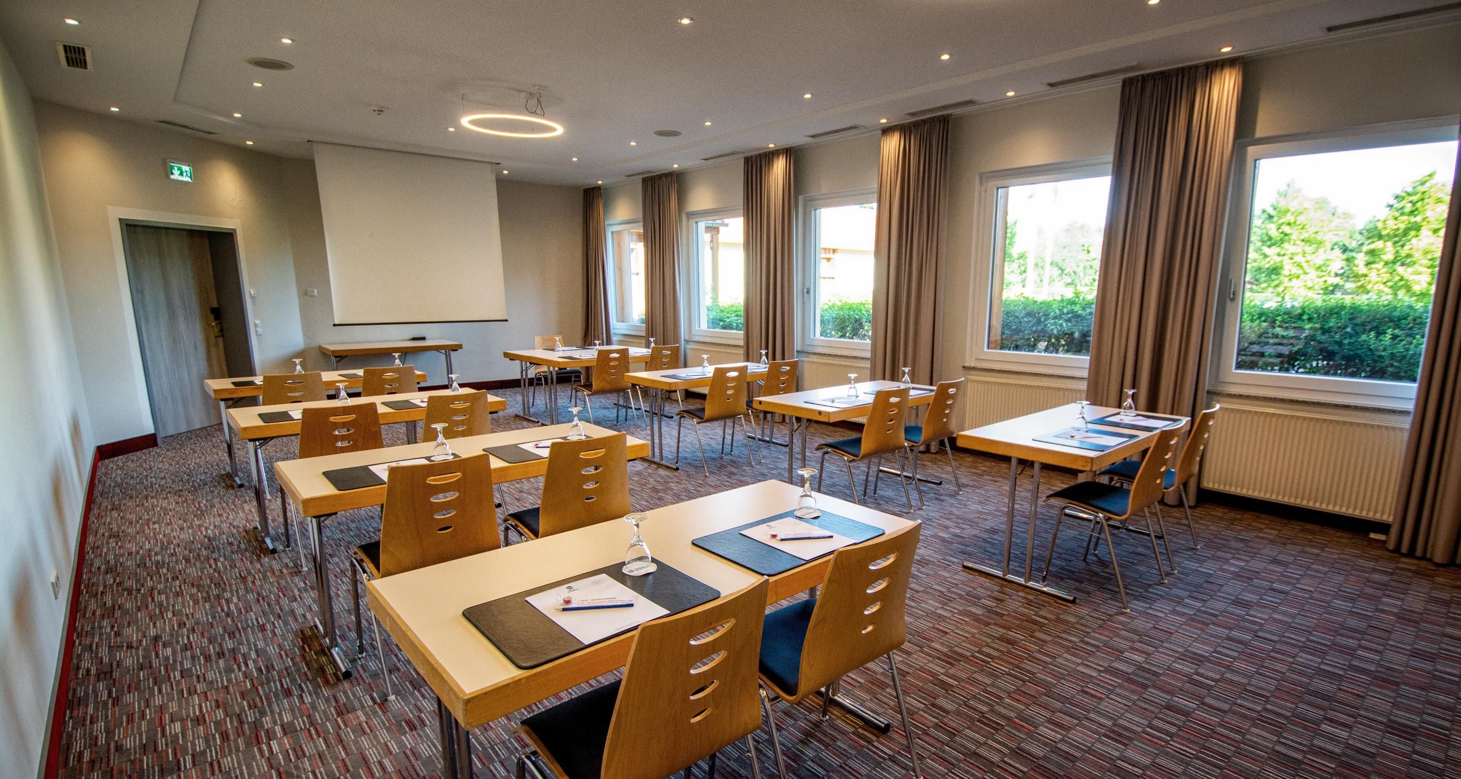 Une salle de conférence équipée de plusieurs tables et chaises, de grandes fenêtres avec des rideaux et d’un écran de projection.