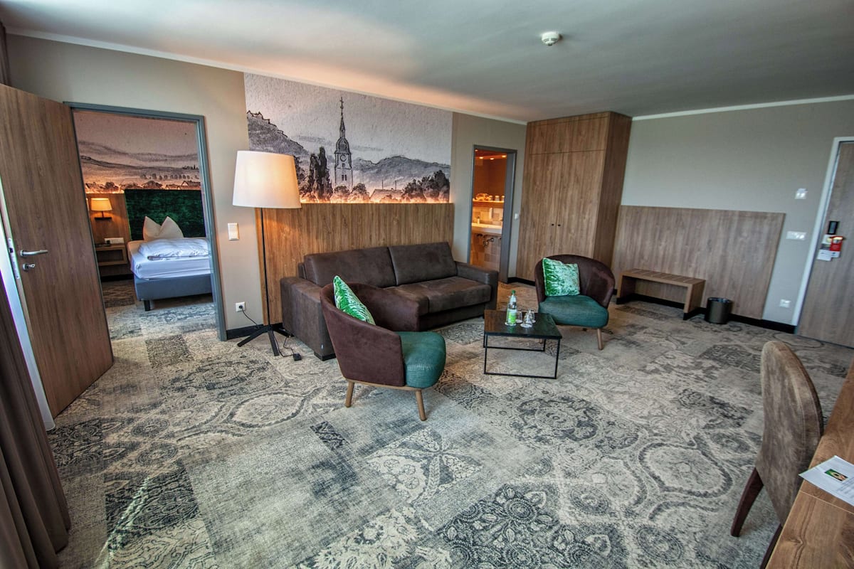 A hotel room with a large bed, a sofa, a chair, a coffee table, and a painting of a cityscape.