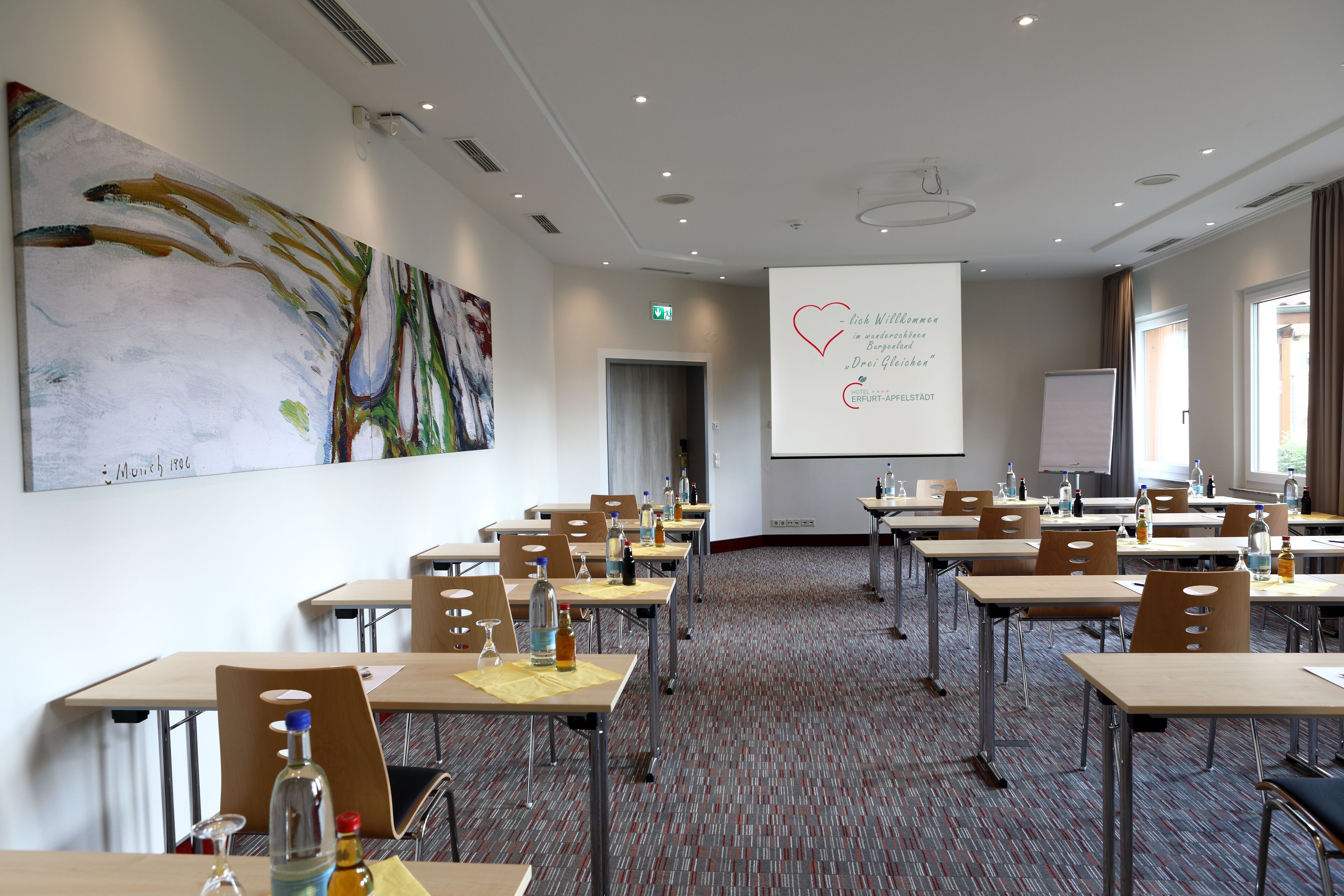A conference room with tables, chairs, a projector screen, and a large abstract painting on the wall.