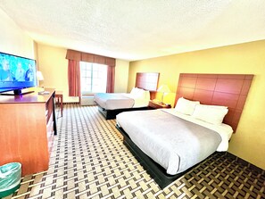 A hotel room with a bed, a desk with a television, a chair, a bedside table with a lamp, and a window with curtains.