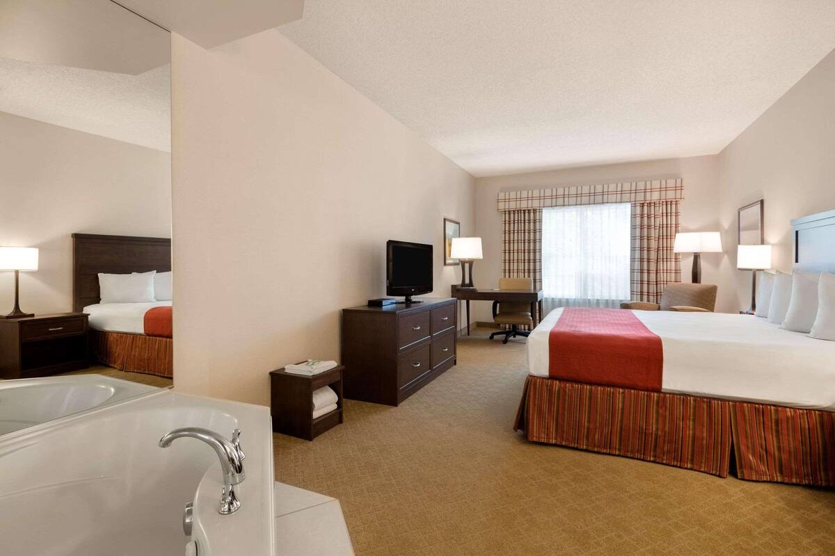 A hotel room with a large bed, a desk, a television, a desk lamp, a chair, a nightstand, a lamp, a window with curtains, and a bathroom with a bathtub and sink.