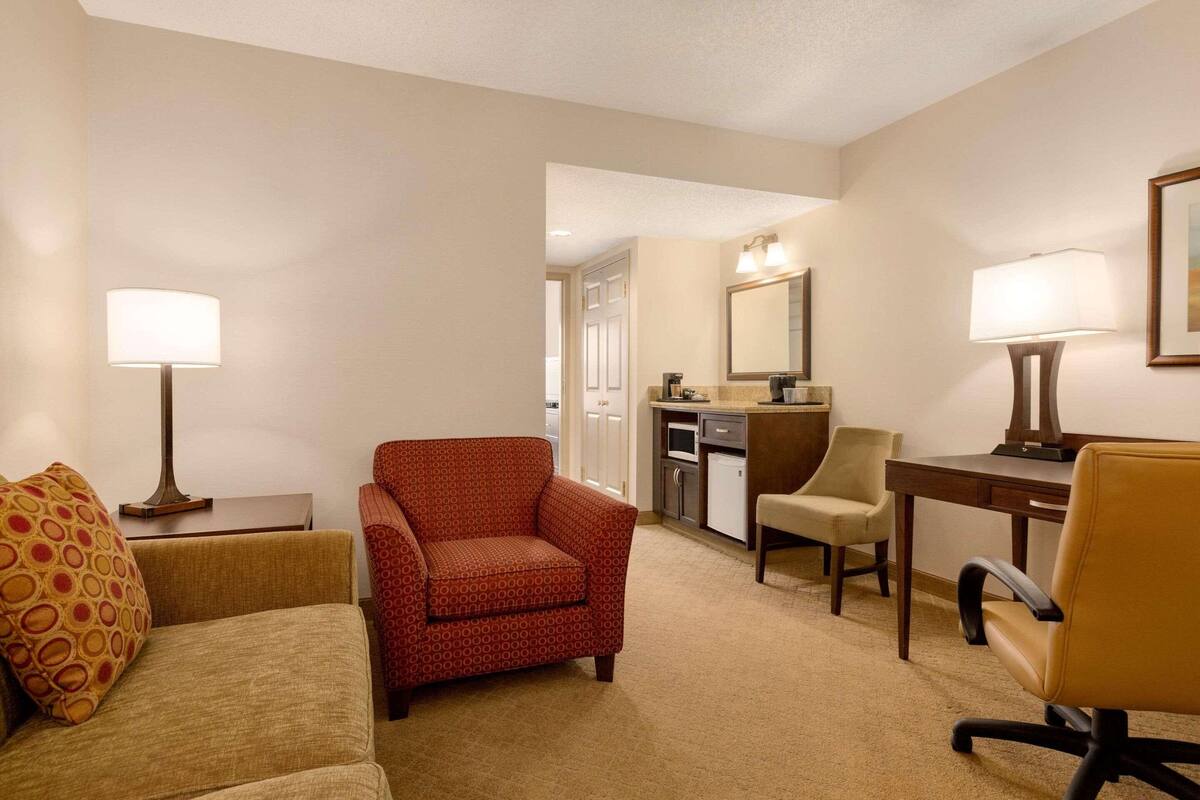 A hotel room with a sofa, armchair, desk, and kitchenette.