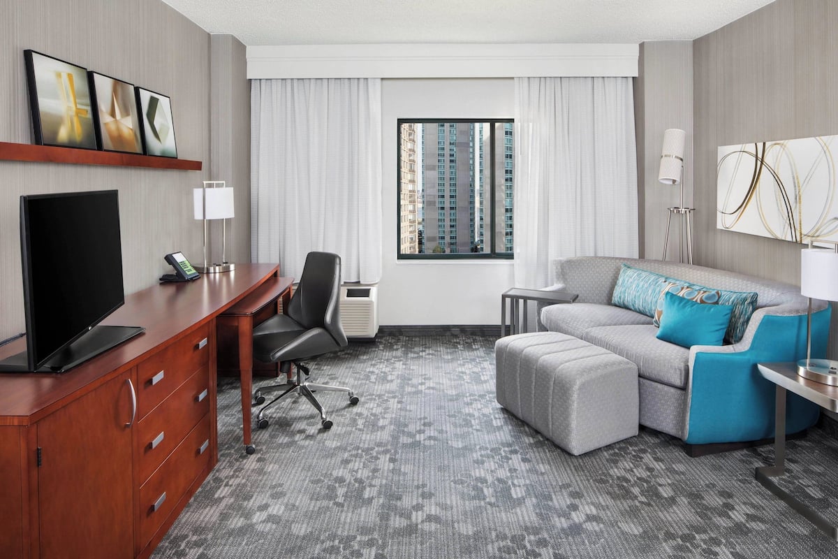 Une chambre d’hôtel équipée d’une télévision à écran plat, d’un bureau avec une chaise, d’un canapé gris avec des coussins bleus, d’une petite table d’appoint et d’une fenêtre donnant sur la ville.