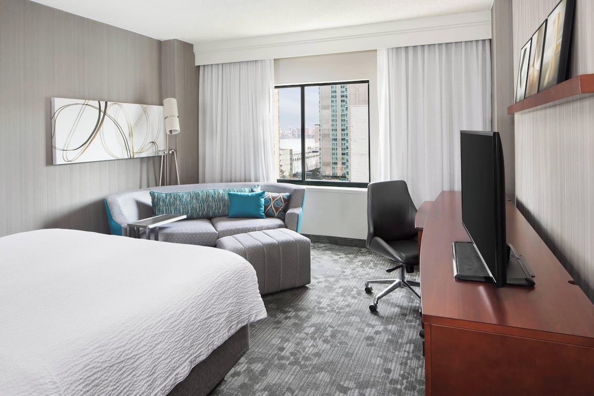 Une chambre d’hôtel avec un grand lit, un canapé, un bureau avec une télévision, une lampe et une fenêtre donnant sur la ville.
