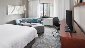 A hotel room with a large bed, a sofa, a desk with a TV, a lamp, and a window with a city view.