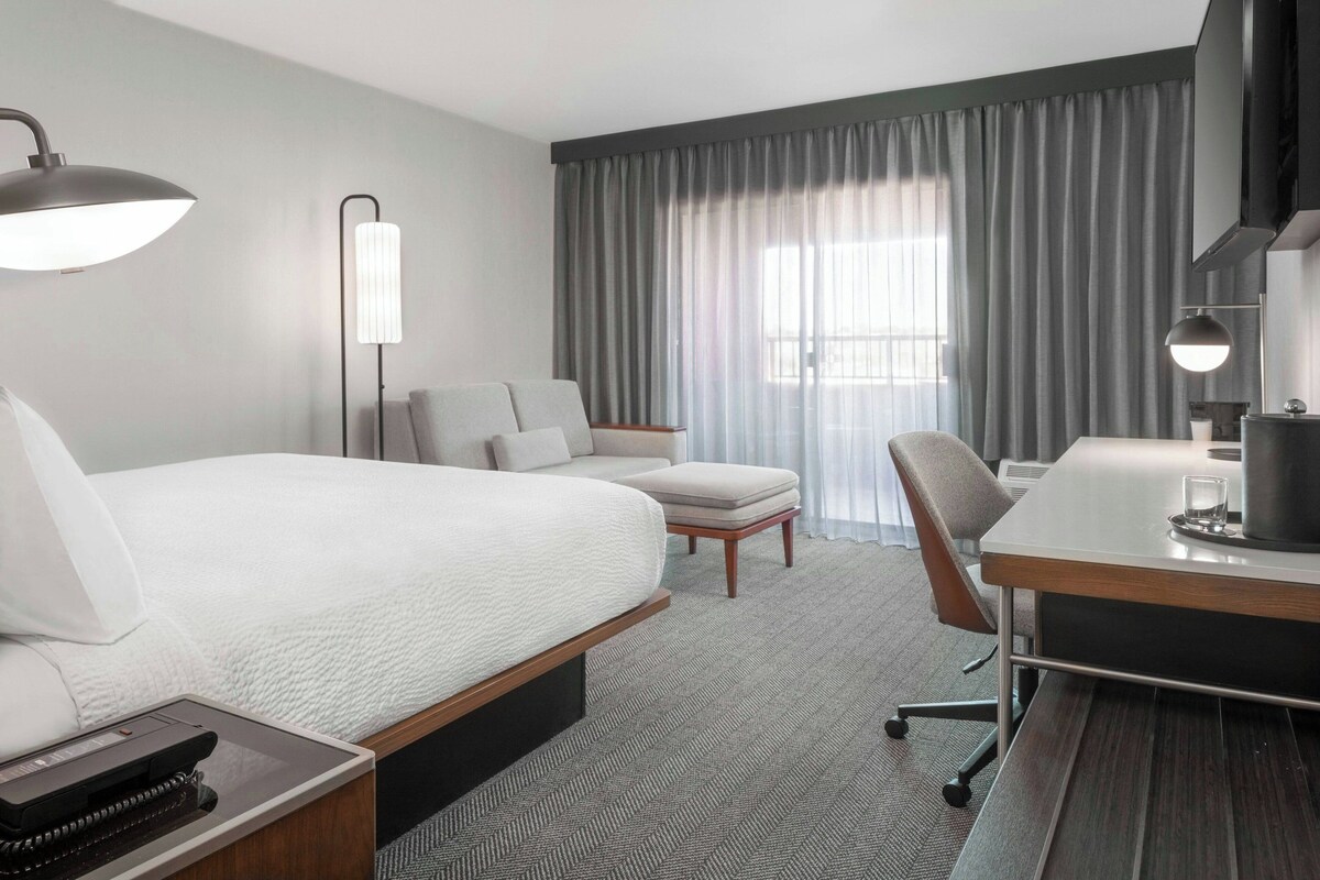 A hotel room with a large bed, a desk, a chair, a lamp, and a TV.