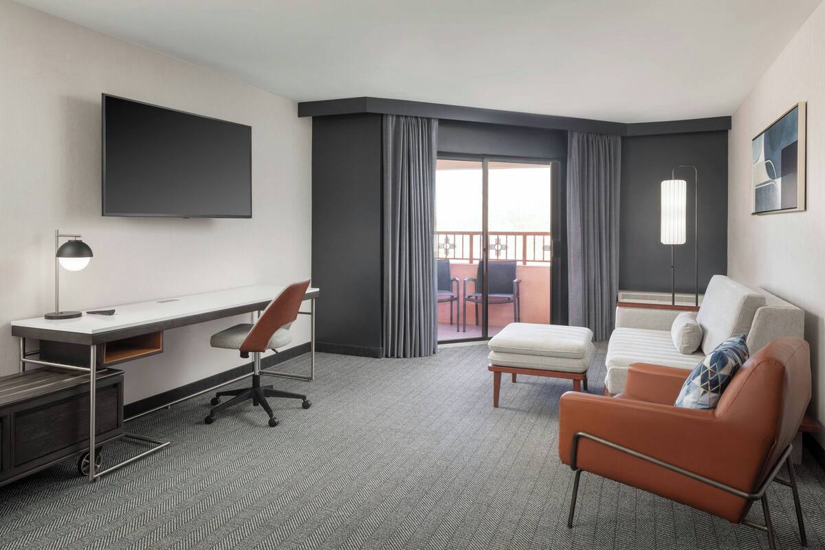 A hotel room with a flat-screen TV, a desk with a chair, a sofa, and a balcony with a view.