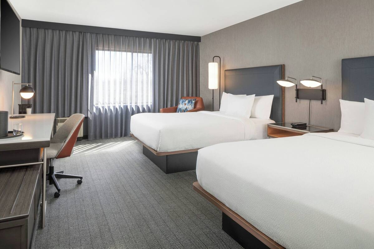 A hotel room with two beds, a desk, a chair, and a large window with curtains.