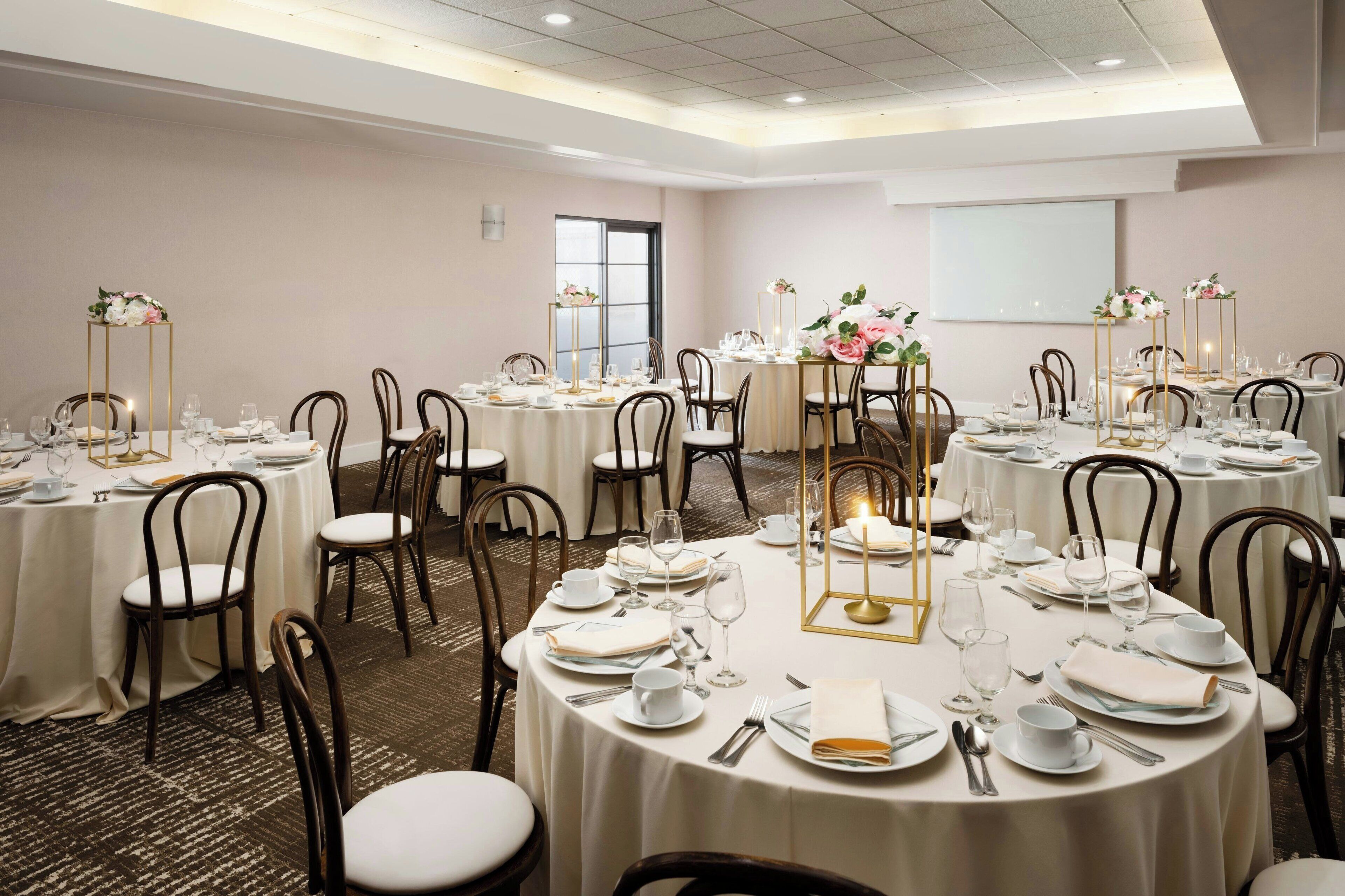 A conference room with round tables, white tablecloths, and chairs. Each table is set with plates, glasses, and cutlery.