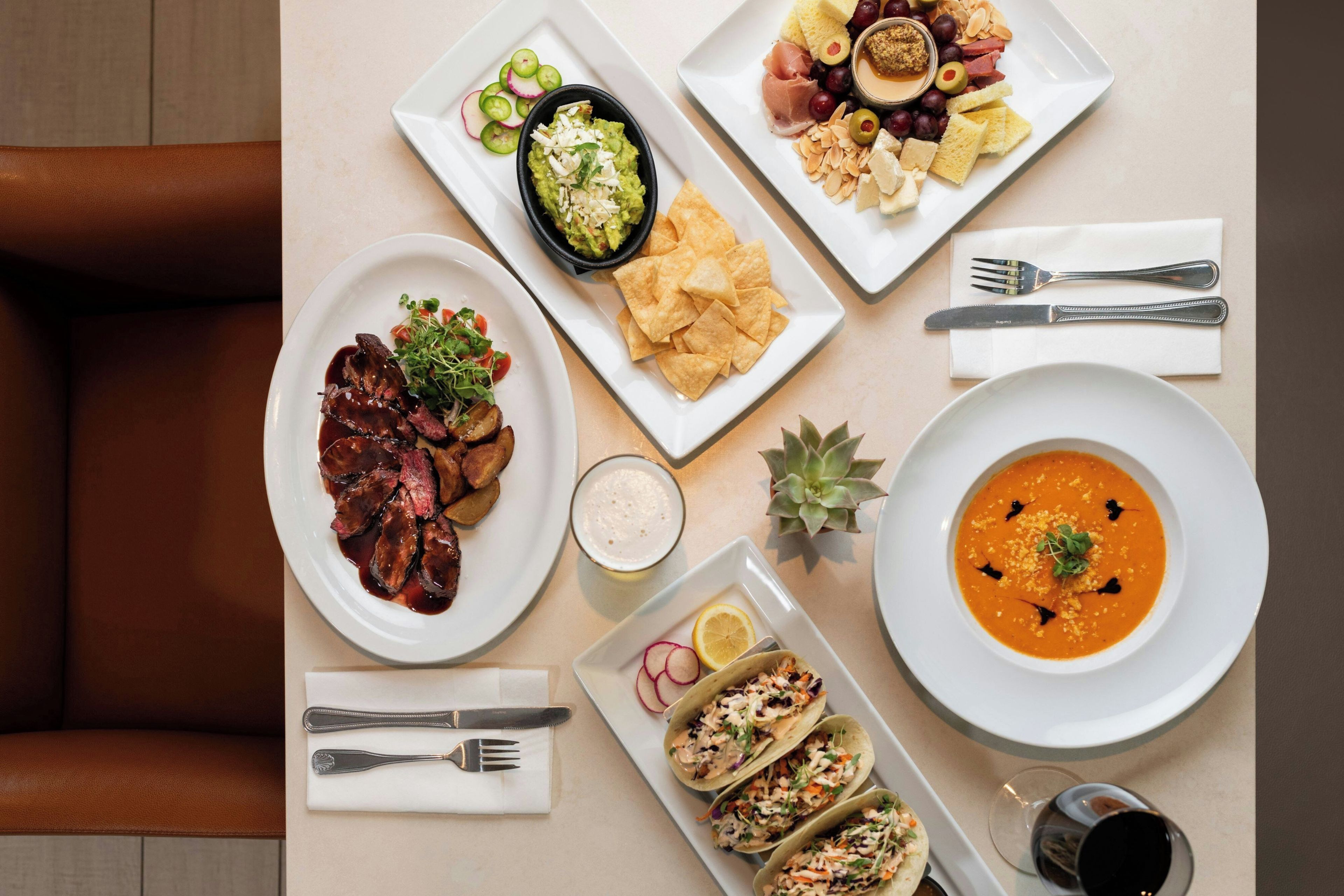 A table with various dishes including steak, soup, and tacos, along with utensils and a small plant.