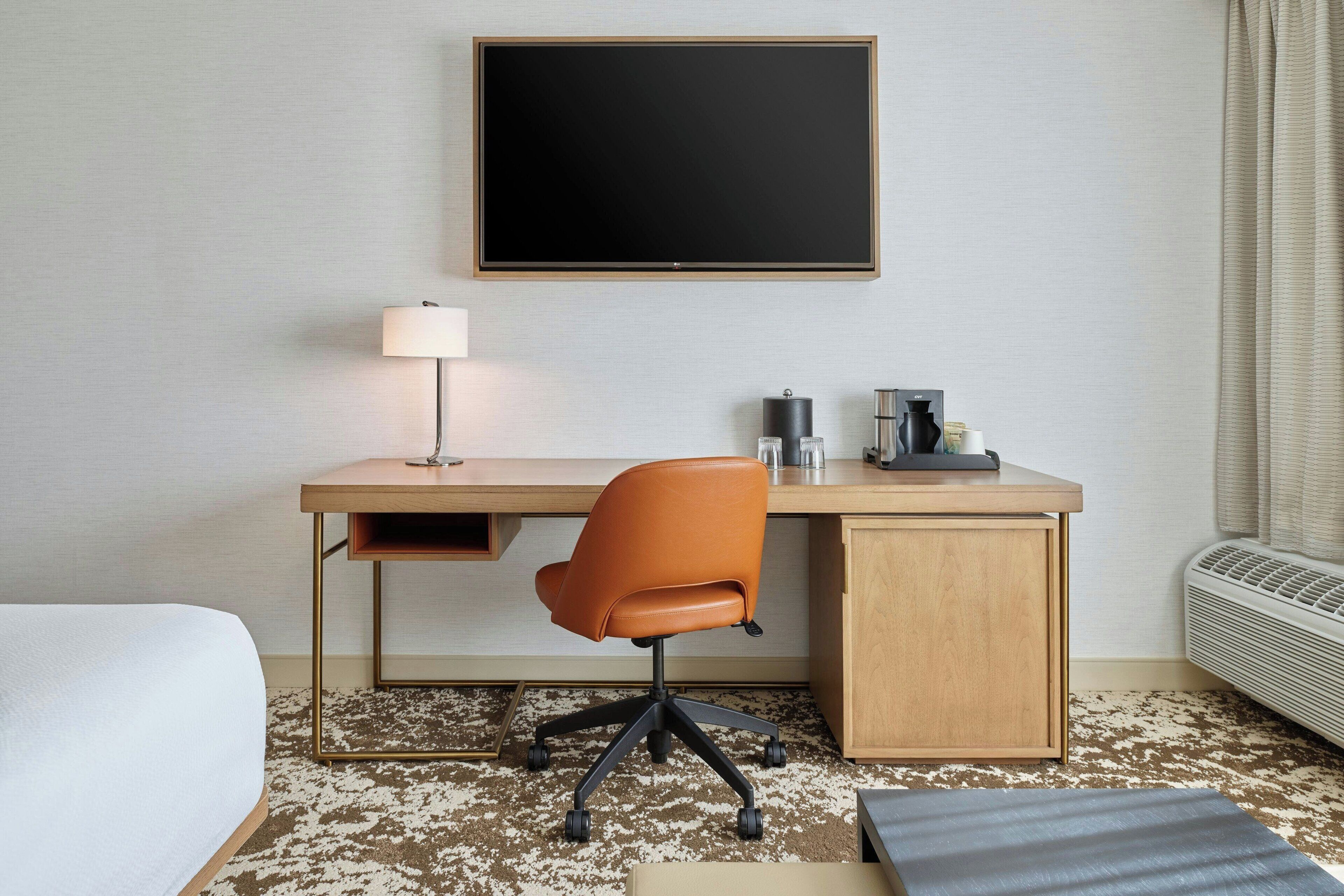 A hotel room with a bed, a desk with a chair, a flat-screen TV, a lamp, a coffee maker, and an air conditioner.