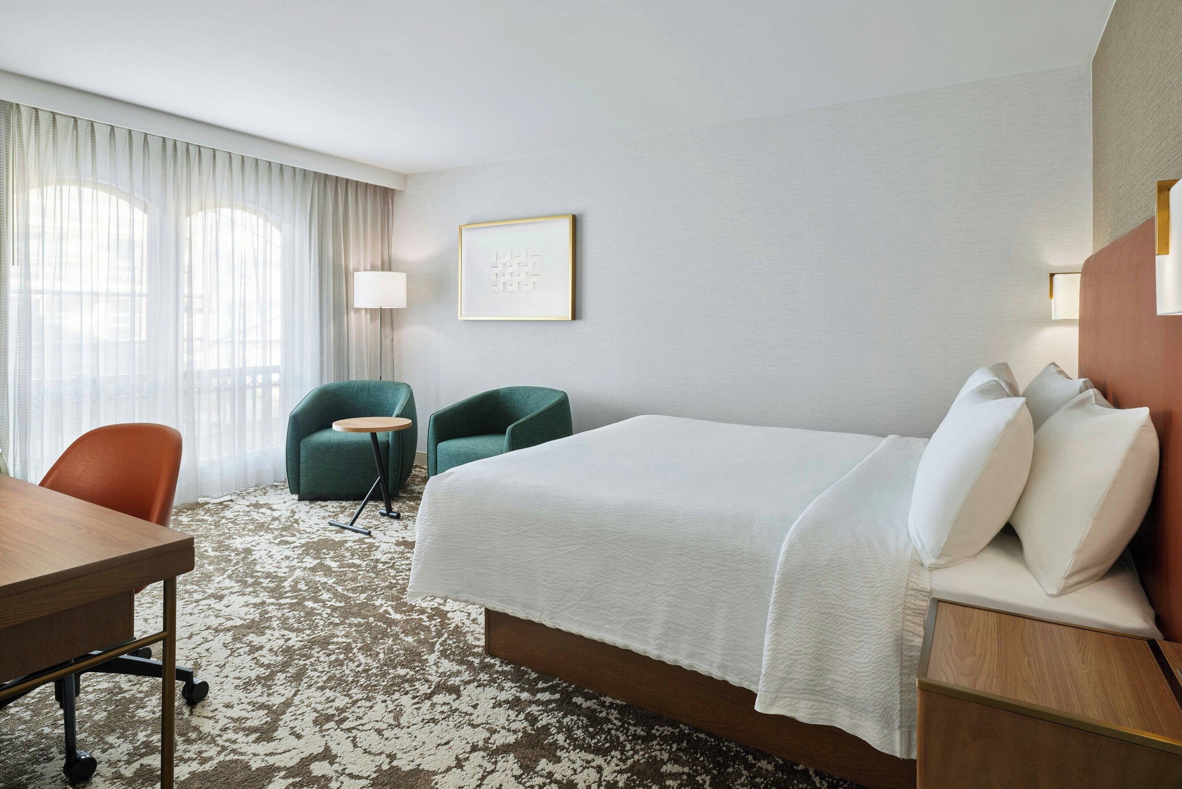 A hotel room with a large bed, a desk, two chairs, a small table, and a lamp.