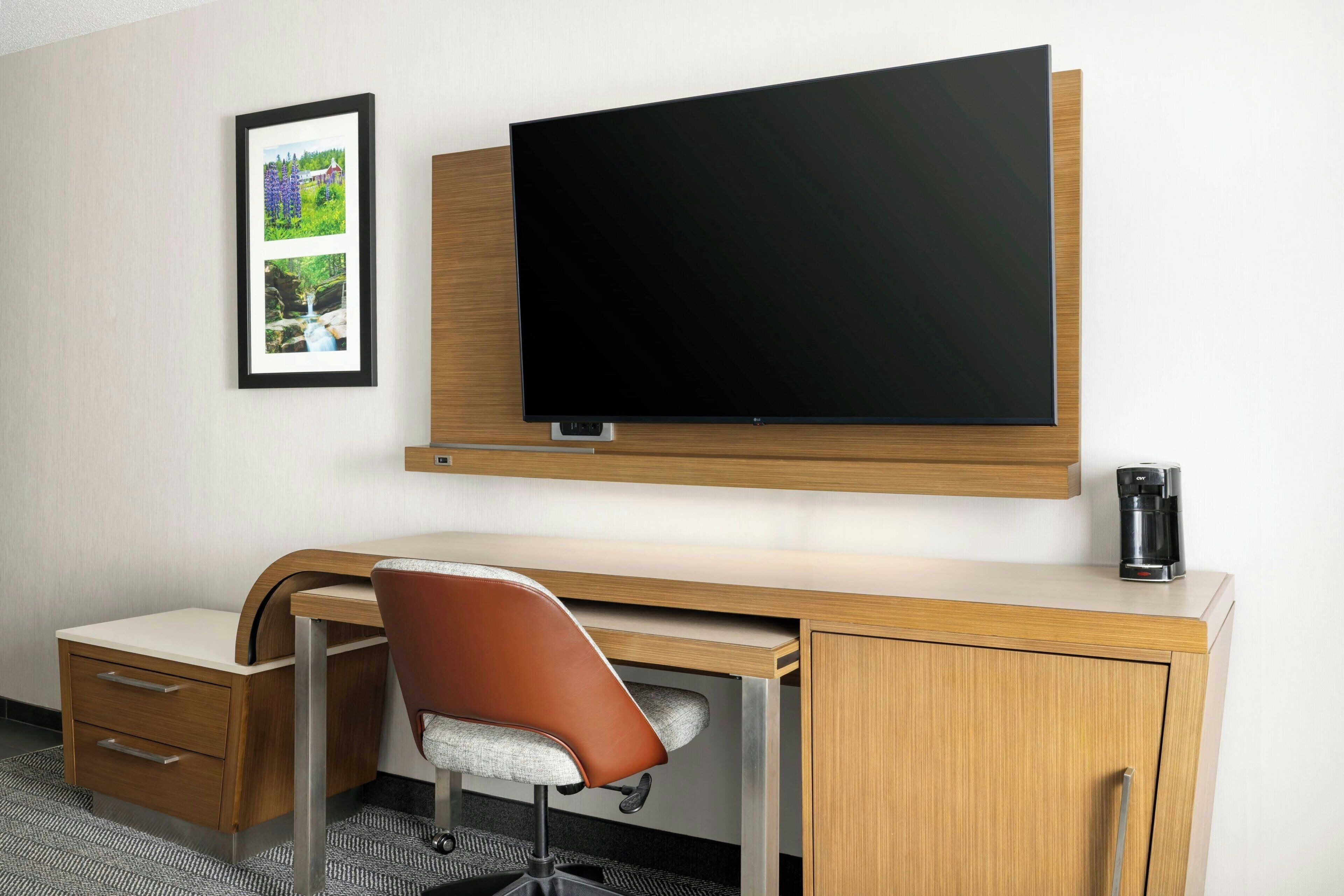 A hotel room with a flat-screen TV mounted on the wall, a wooden desk with a chair, a small cabinet, and a framed picture on the wall.