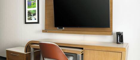 Habitación de hotel con televisor de pantalla plana montado en la pared, un escritorio de madera con una silla, un mueble pequeño y una fotografía en el marco.