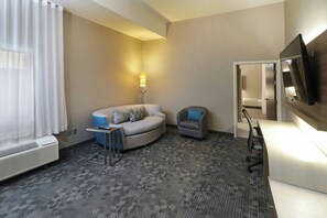A hotel room with a sofa, armchair, desk, TV, and a lamp.