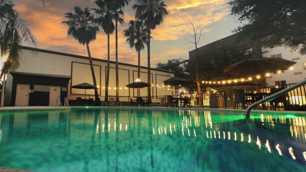 Piscina com palmeiras e guarda-sóis, cercada por edifícios e luzes de corda.