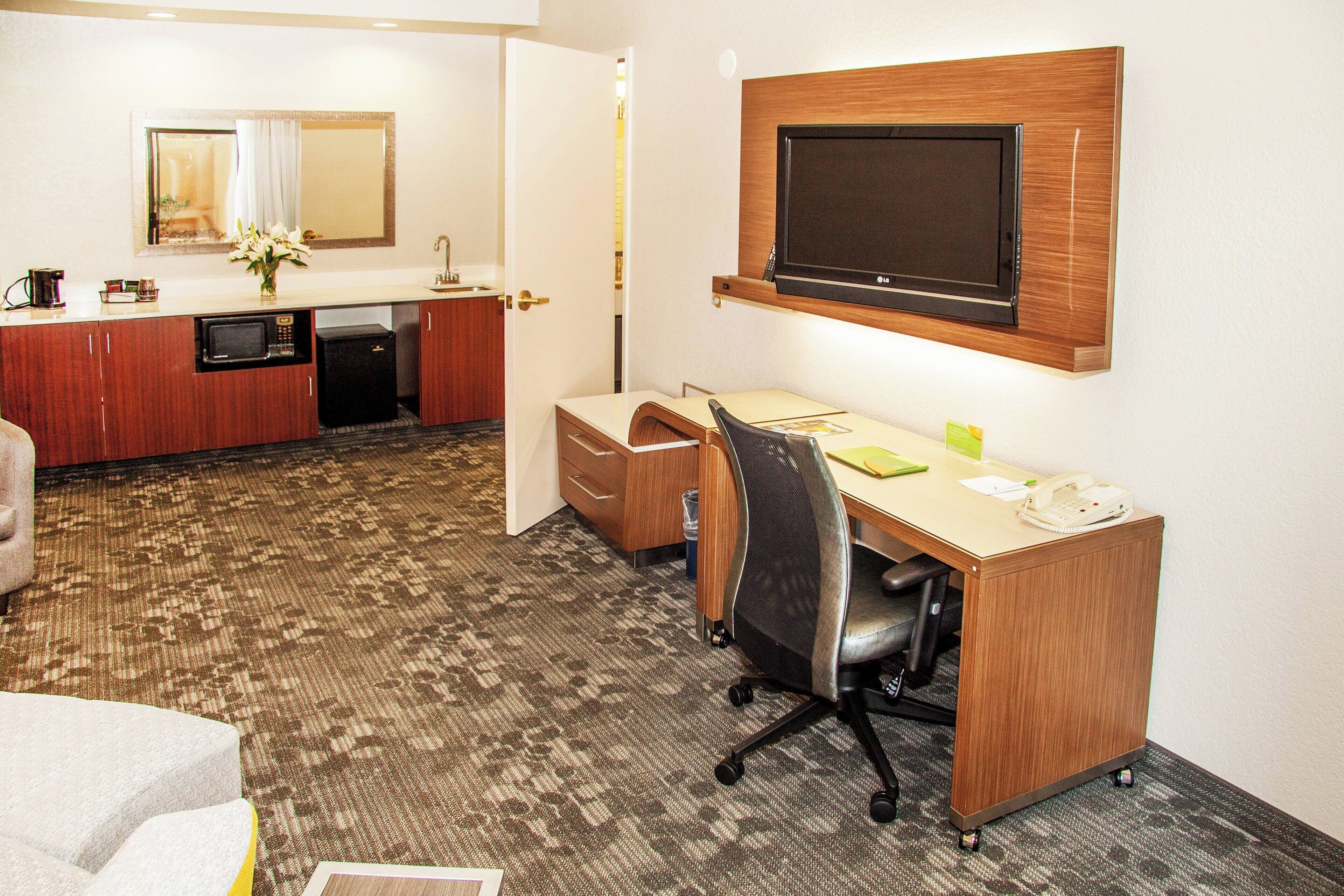 A hotel room with a desk, chair, microwave, and a television mounted on the wall.