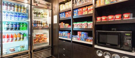 Un área de comida autoservicio con dispensadores refrigerados para bebidas, estantes de bocadillos y un microondas.