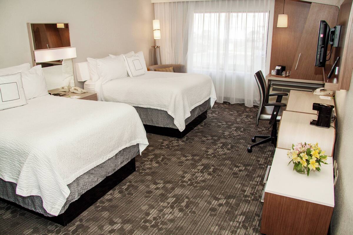 Une chambre d’hôtel avec deux lits, un bureau, une chaise, une lampe de bureau, un téléphone et un petit bureau agrémenté d’un vase de fleurs.