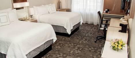 A hotel room with two beds, a desk, a chair, a desk lamp, a telephone, and a small desk with a vase of flowers.