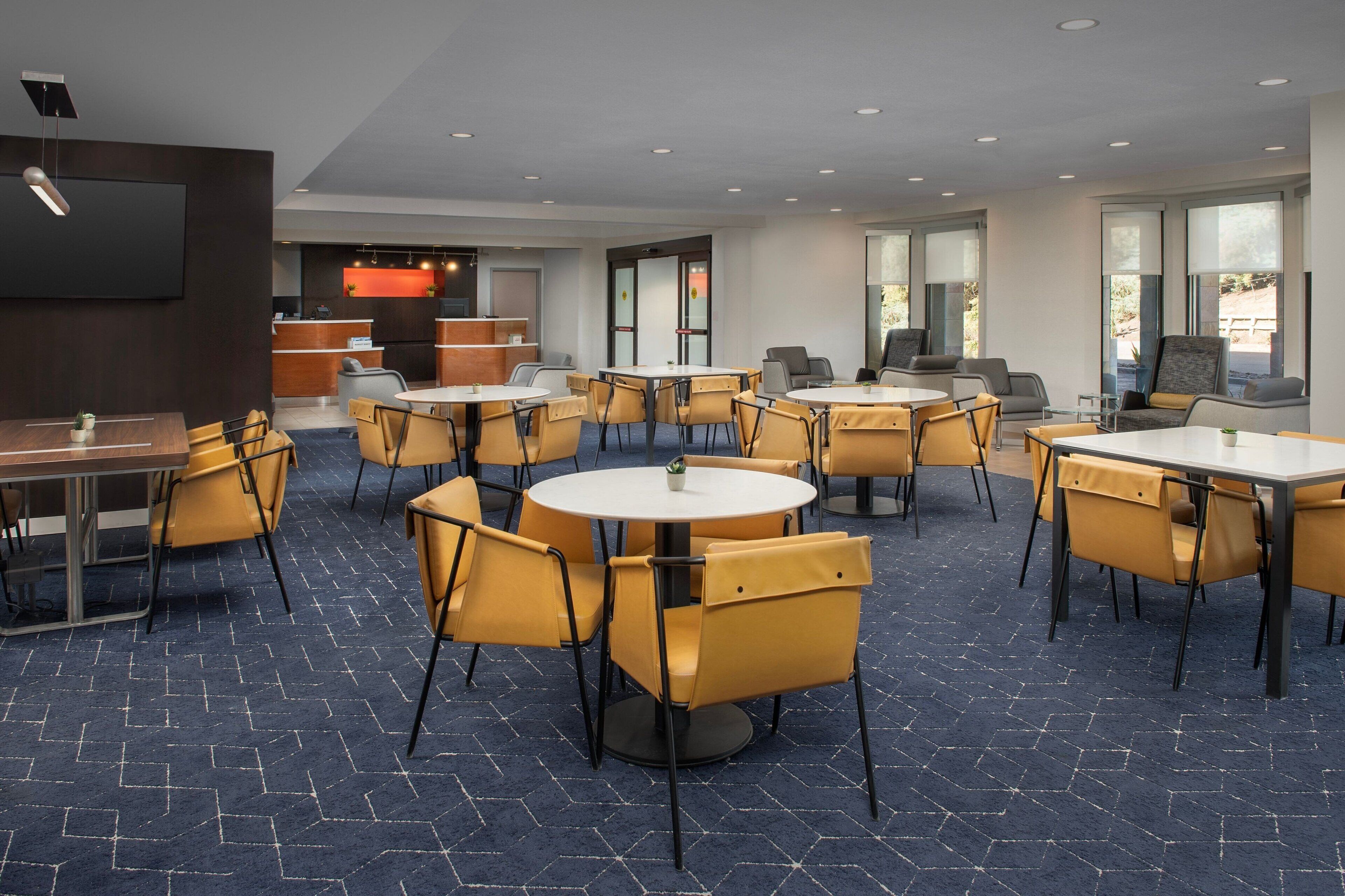 A modern hotel lobby with round tables and yellow chairs, a reception desk, and large windows.