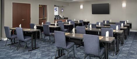 A conference room with multiple tables and chairs, a flat-screen TV mounted on the wall, and a kitchenette area.
