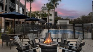 An outdoor seating area with a fire pit, lounge chairs, and umbrellas, overlooking a swimming pool and a building with multiple balconies.