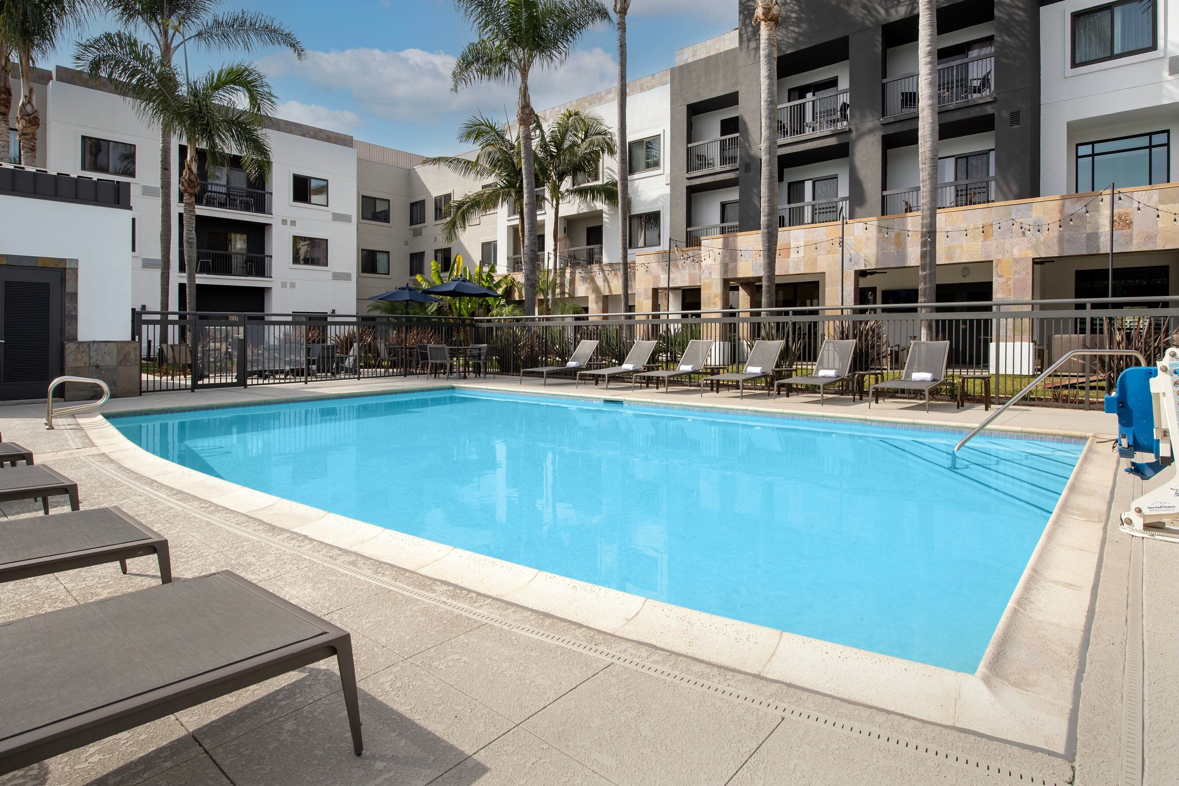Piscina com espreguiçadeiras e palmeiras, rodeada por um edifício com varandas.