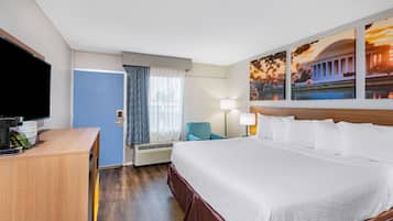 A hotel room with a large bed, a desk with a flat-screen TV, a chair, a lamp, and a window with curtains.