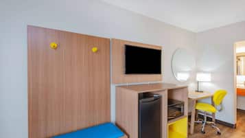 A compact hotel room with a wooden wardrobe, a flat-screen TV, a small refrigerator, a desk with a chair, and a mirror.