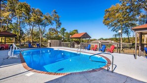 A swimming pool with a brick edge, surrounded by a wooden deck, and a covered seating area with Adirondack chairs. - Motel 6 Hot Springs, AR (Hot Springs)