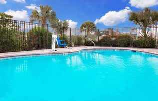 Una piscina azul cristalina con una cerca metálica, una silla de descanso azul y palmeras al fondo.