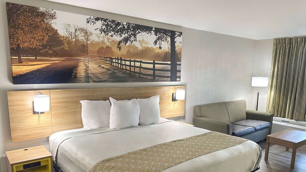 A hotel room with a large bed, a sofa, a small table, a nightstand, and a wall-mounted lamp. - Days Inn & Suites by Wyndham Opelousas (Opelousas)