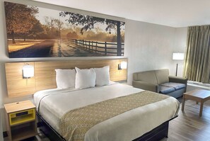 A hotel room with a large bed, a sofa, a small table, a nightstand, and a wall-mounted lamp. - Days Inn & Suites by Wyndham Opelousas (Opelousas)