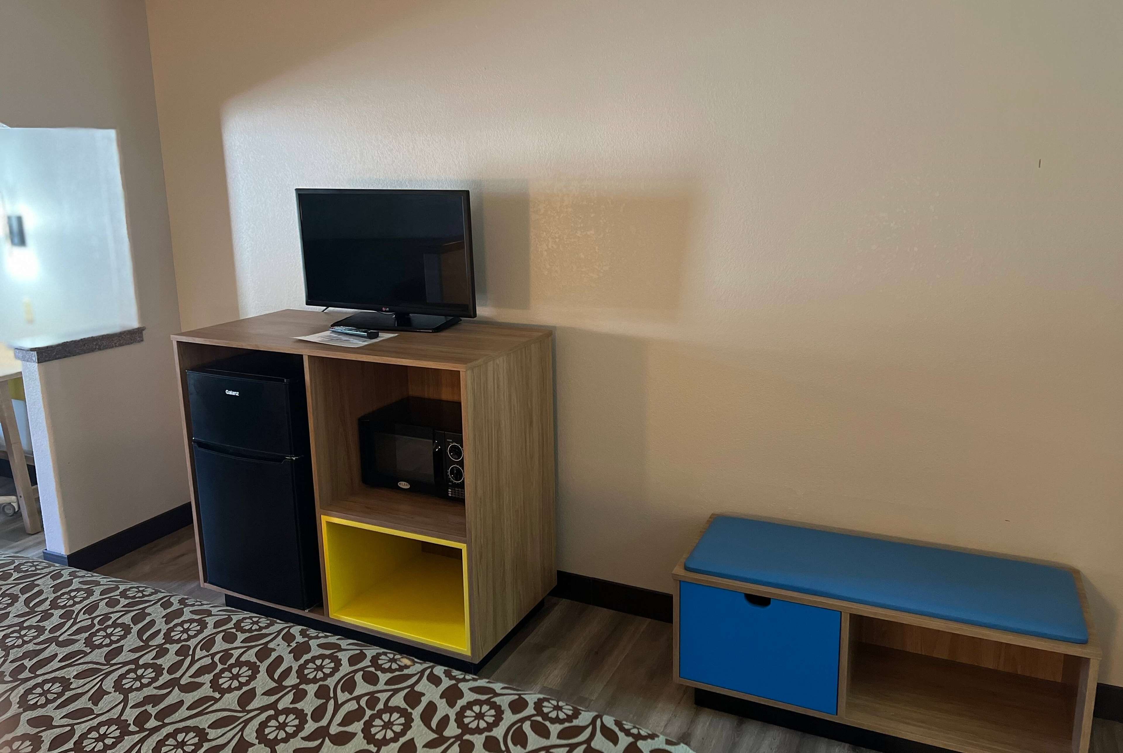 Une petite pièce comprenant un lit, un bureau avec un ordinateur, une télévision et une petite armoire bleue.