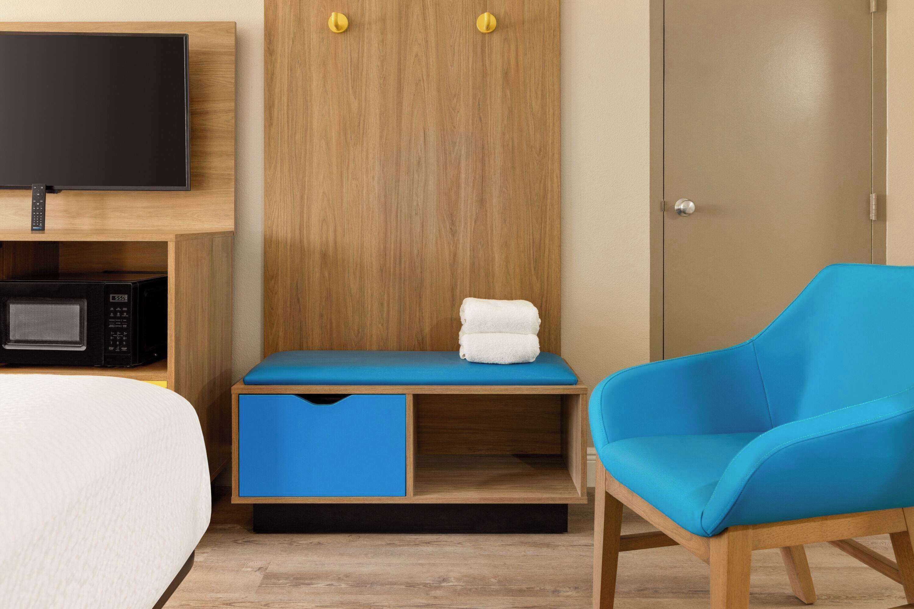 Habitación de hotel con cama, televisión montada en la pared, microondas, armario de madera con dos puertas, una silla azul y un pequeño banco con una toalla.