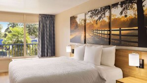 Une chambre d’hôtel avec un grand lit, une table de chevet, une lampe fixée au mur et, par la fenêtre, une vue sur des palmiers et une clôture.