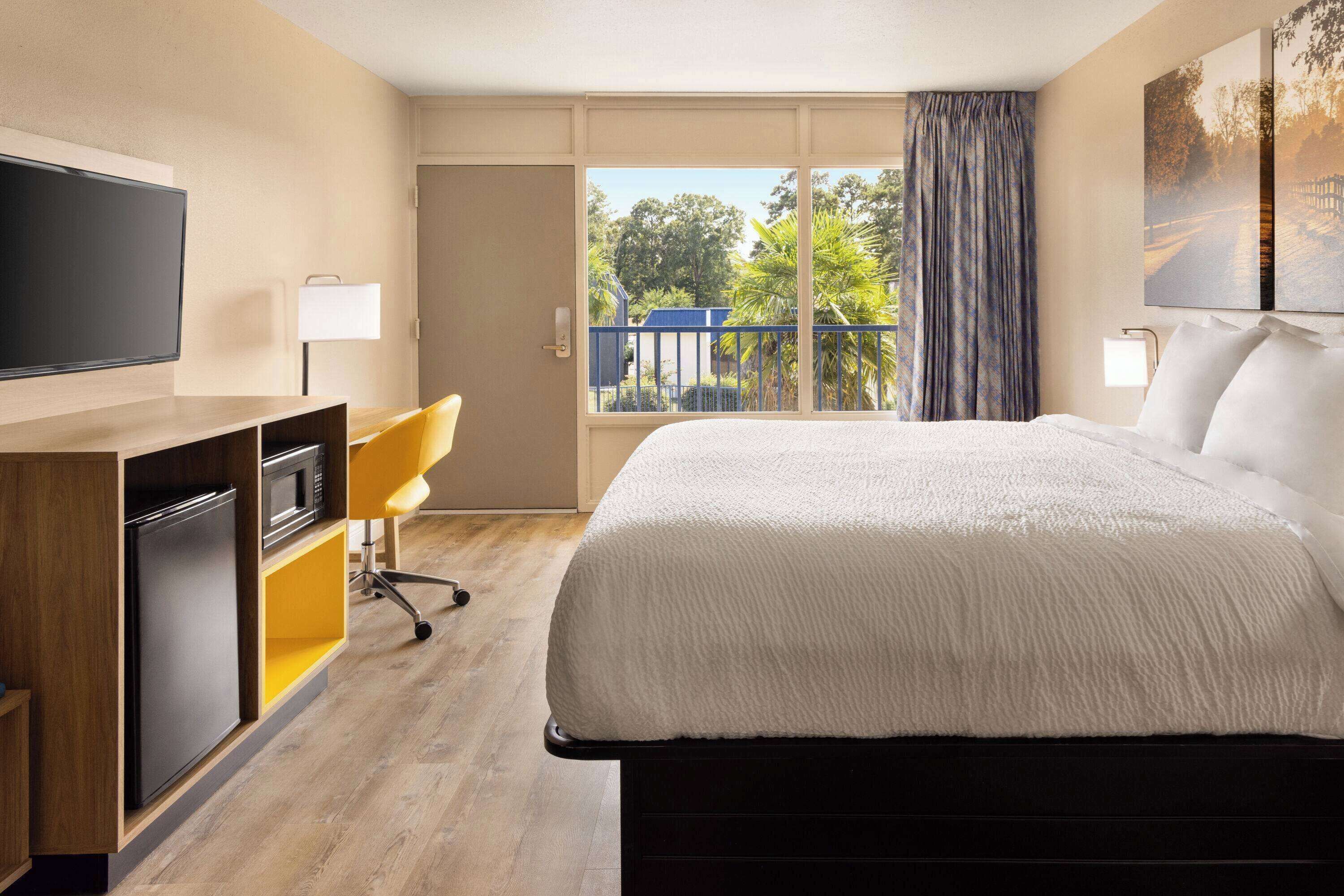 A hotel room with a bed, a TV, a desk with a chair, a window with a view of trees, and a balcony.