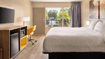Ein Hotelzimmer mit Bett, Fernseher, Schreibtisch mit Stuhl, einem Fenster mit Blick auf Bäume und einem Balkon.