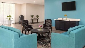 Un hall d’hôtel avec des canapés de couleur turquoise, une table ronde ornée d’un vase de fleurs, deux fauteuils, un buffet en bois foncé, un téléviseur fixé au mur et une plante en pot.