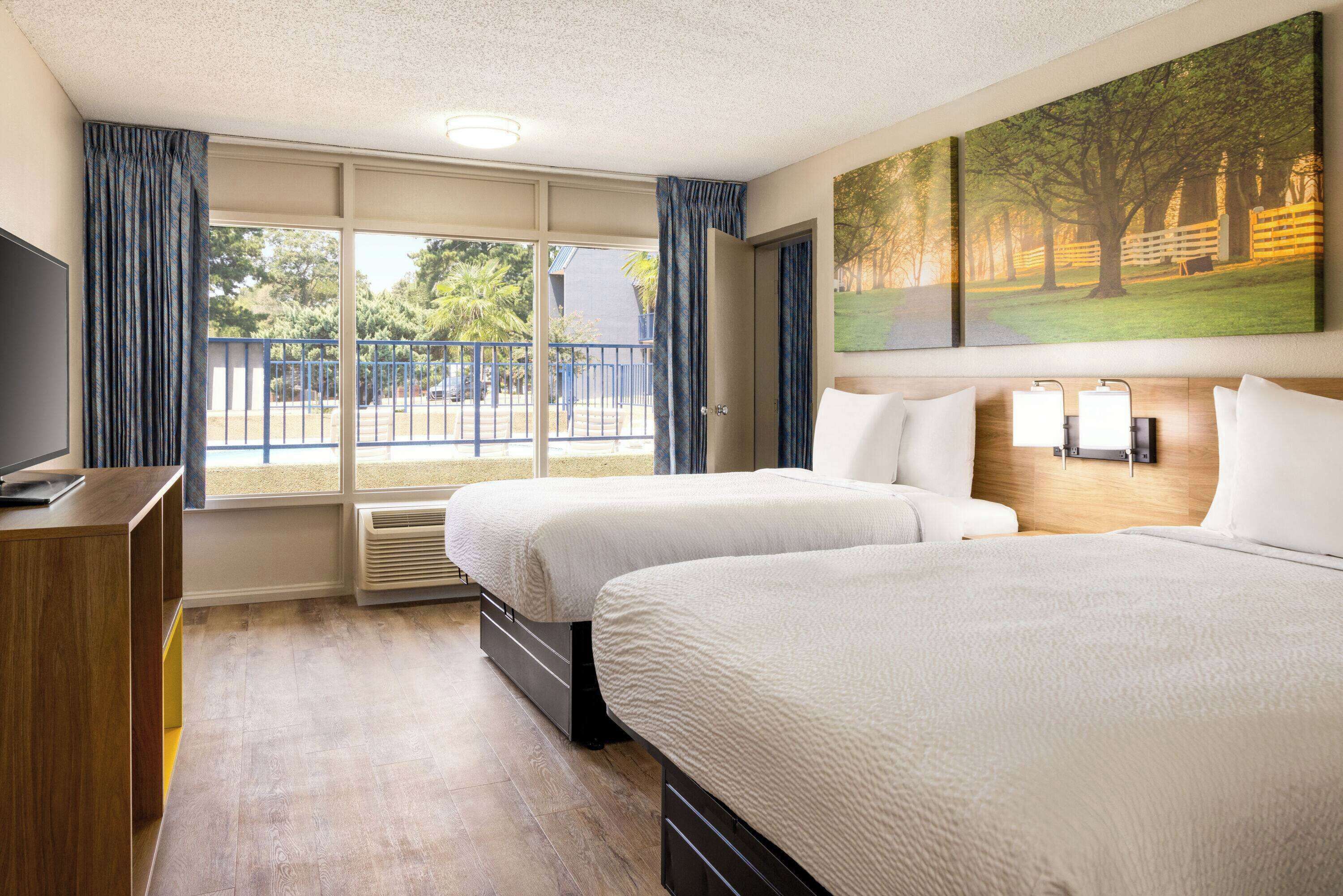Habitación de hotel con dos camas, televisión, un ventanal grande con cortinas y un cuadro en la pared.