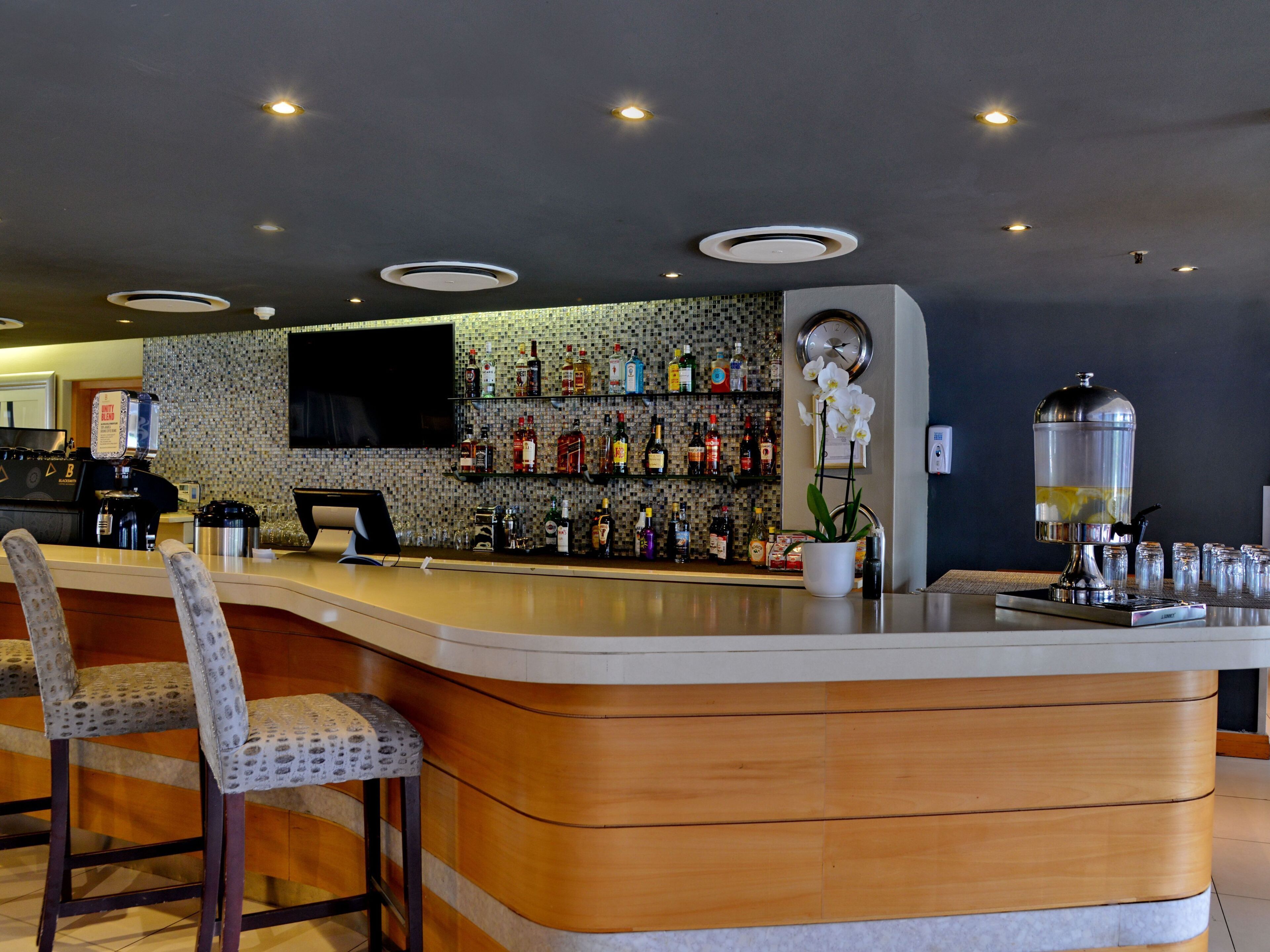 A bar area with a curved counter, bar stools, a television mounted on the wall, and shelves stocked with bottles.