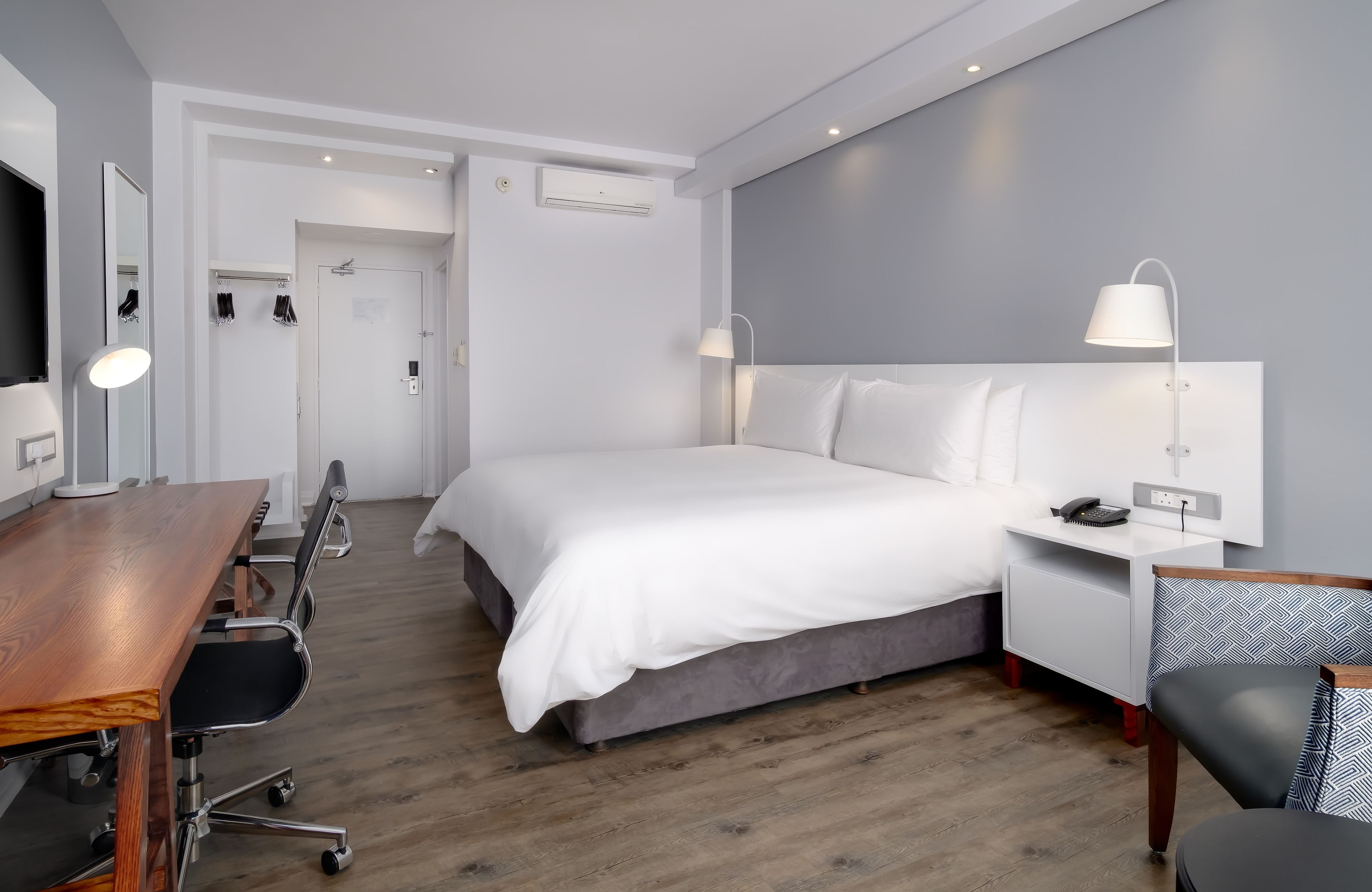 A modern hotel room with a wooden desk, a large bed, a nightstand, and a chair.